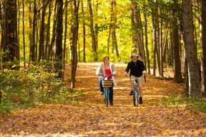 A man and woman ride bikes through the woods on a leaf-covered path in fall on Mackinac Island