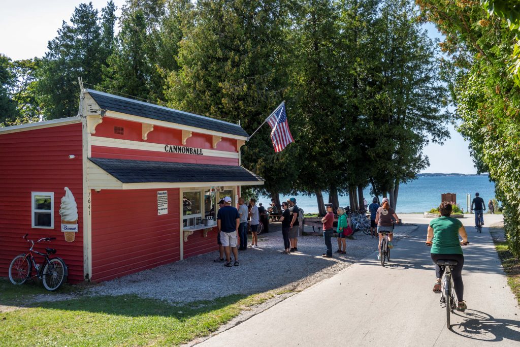 People wait in line to order food at Cannonball Oasis as bicyclists pedal past at Mackinac Island’s British Landing