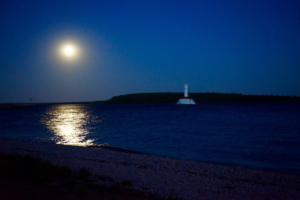 The moon shines on the water off a rocky Mackinac Island beach at night with a lighthouse in the distance