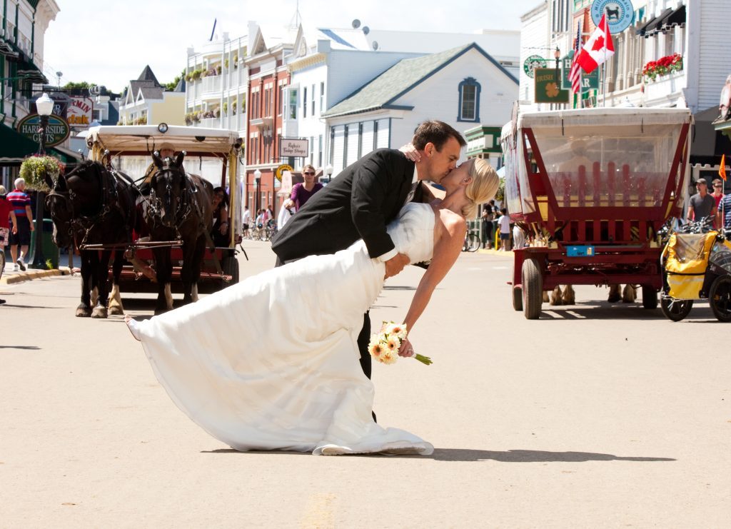 A bride and groom share an iconic Mackinac Island wedding kiss in the middle of Main Street as horse-drawn carriages go past