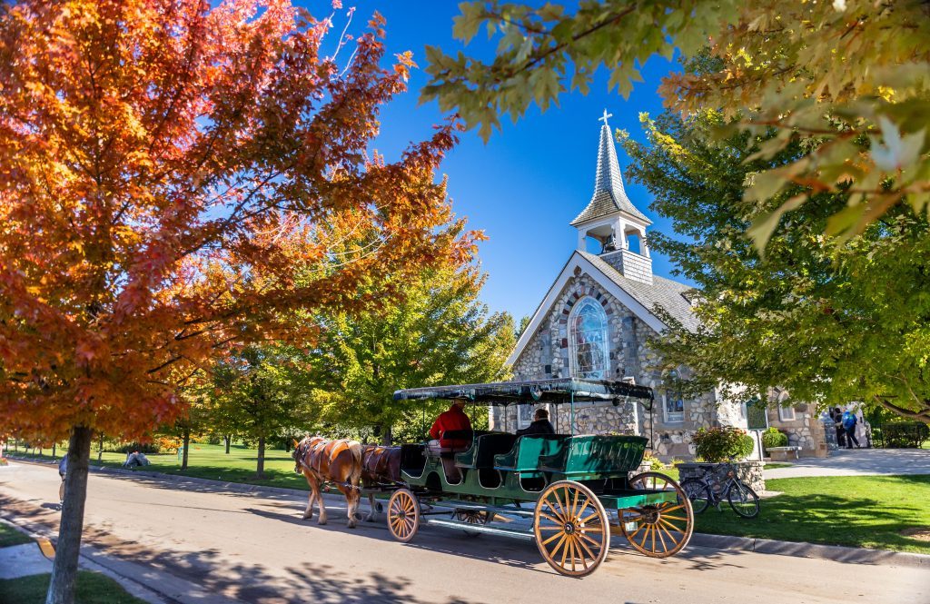 A horse-drawn carriages passes a Mackinac Island church on a sunny fall day with leaves changing colors