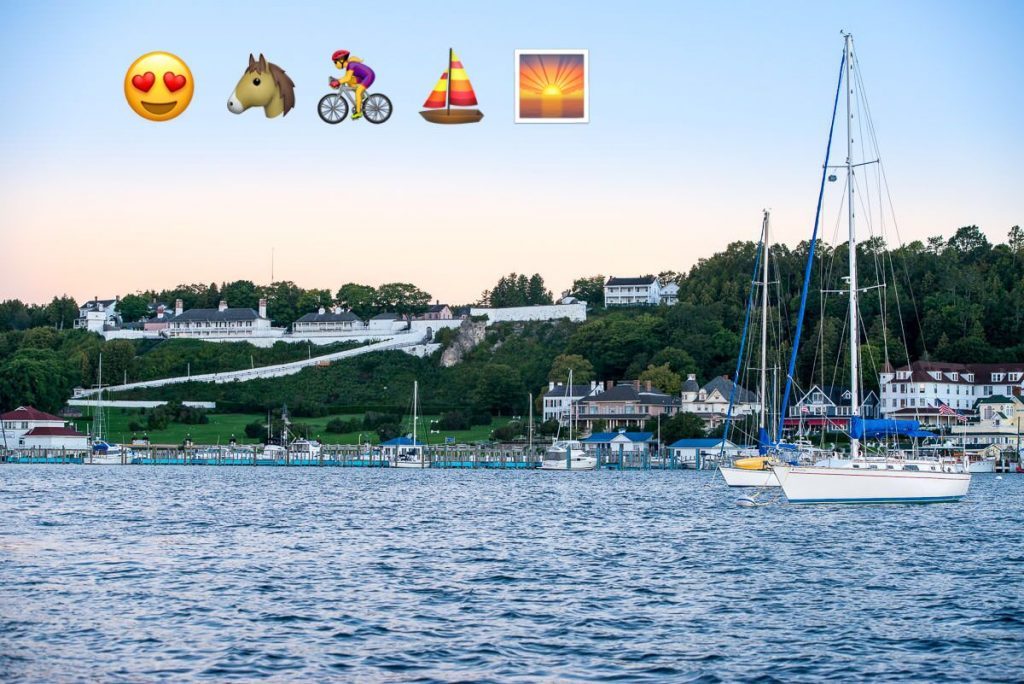 Sailboats in the water by the Mackinac Island marina beneath Fort Mackinac with emojis for happy face, horse, bicycle, boat and sunrise