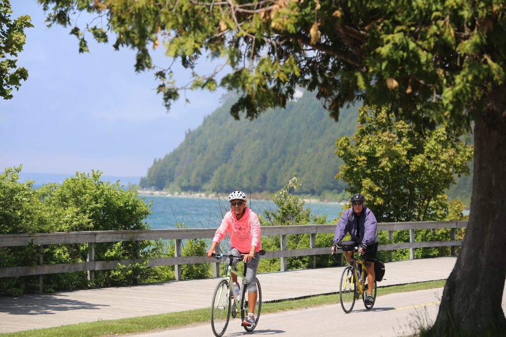 Two women wearing helmets and windbreakers ride bicycles along the Mackinac Island waterfront on a sunny day