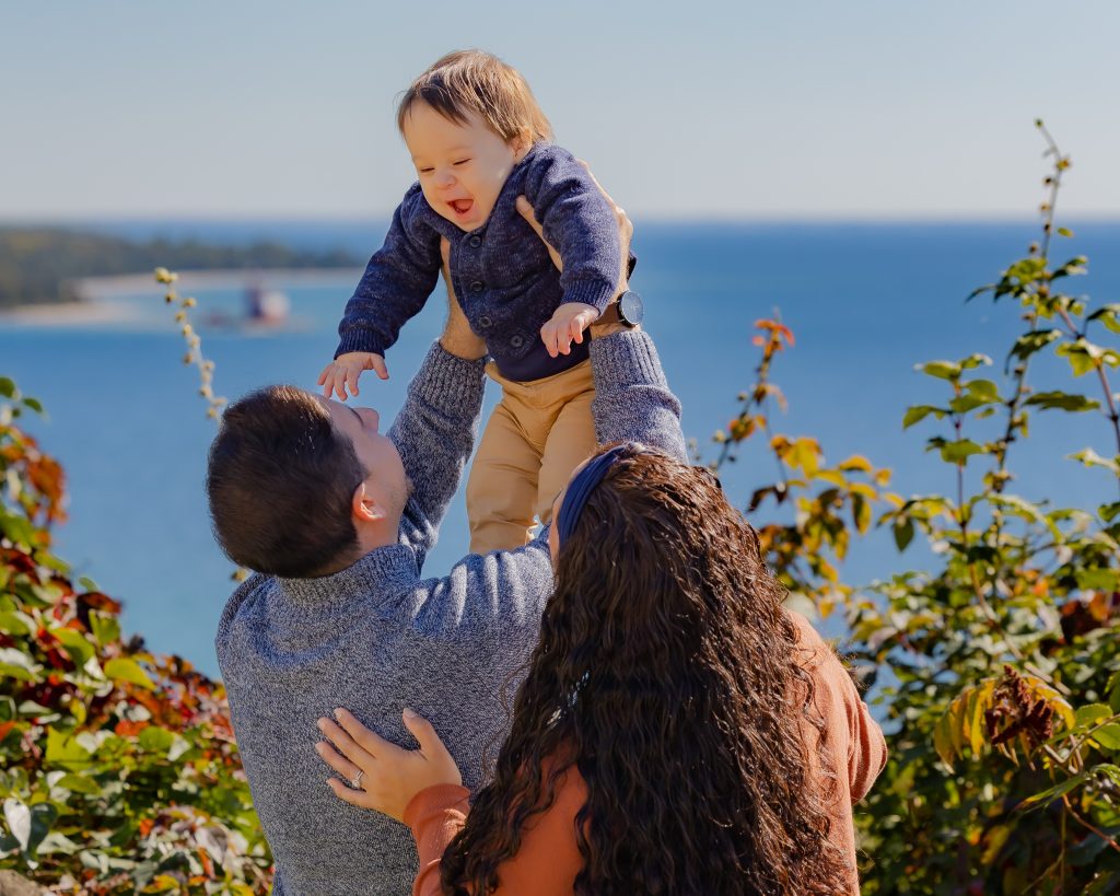A young couple lifts their little boy into the air on Mackinac Island’s West Bluff with lighthouse in the water