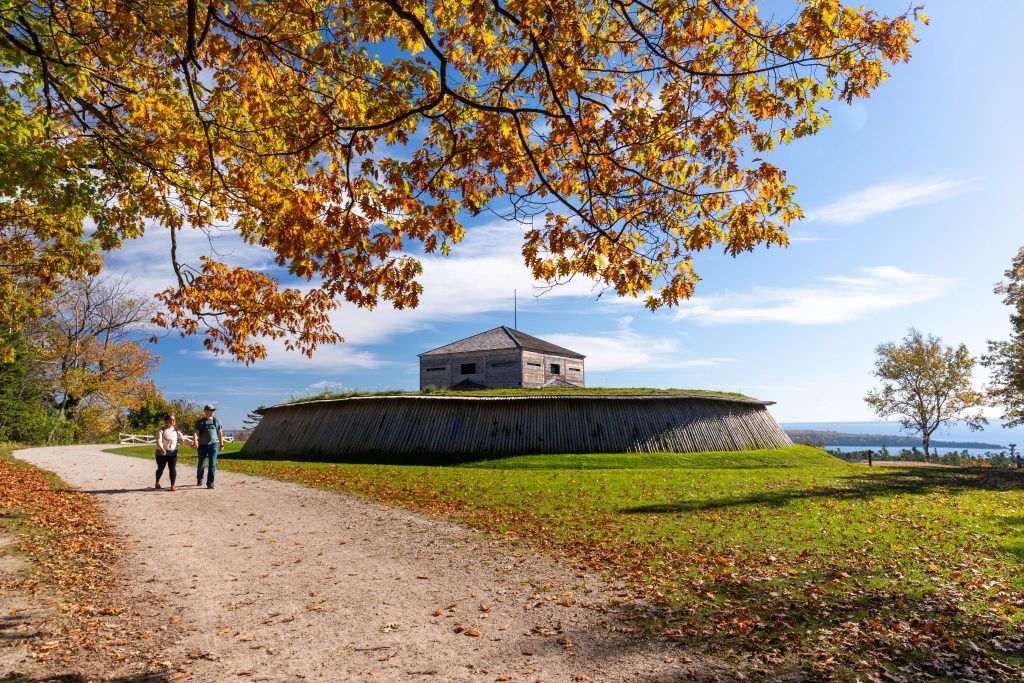 A couple walks hand in hand down a leaf-covered path past Fort Holmes on a sunny fall day on Mackinac Island