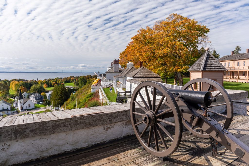 A Fort Mackinac cannon points out over town amid fall colors and blue water with the Mackinac Bridge in background