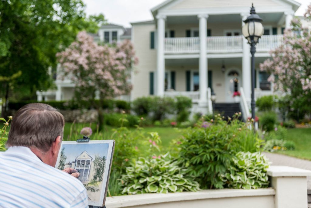 An artist paints the front façade of a Mackinac Island place to stay that can be seen in the background.