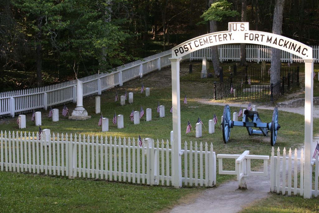 A blue cannon stands guard over the graves inside a white fence in the military Post Cemetery on Mackinac Island