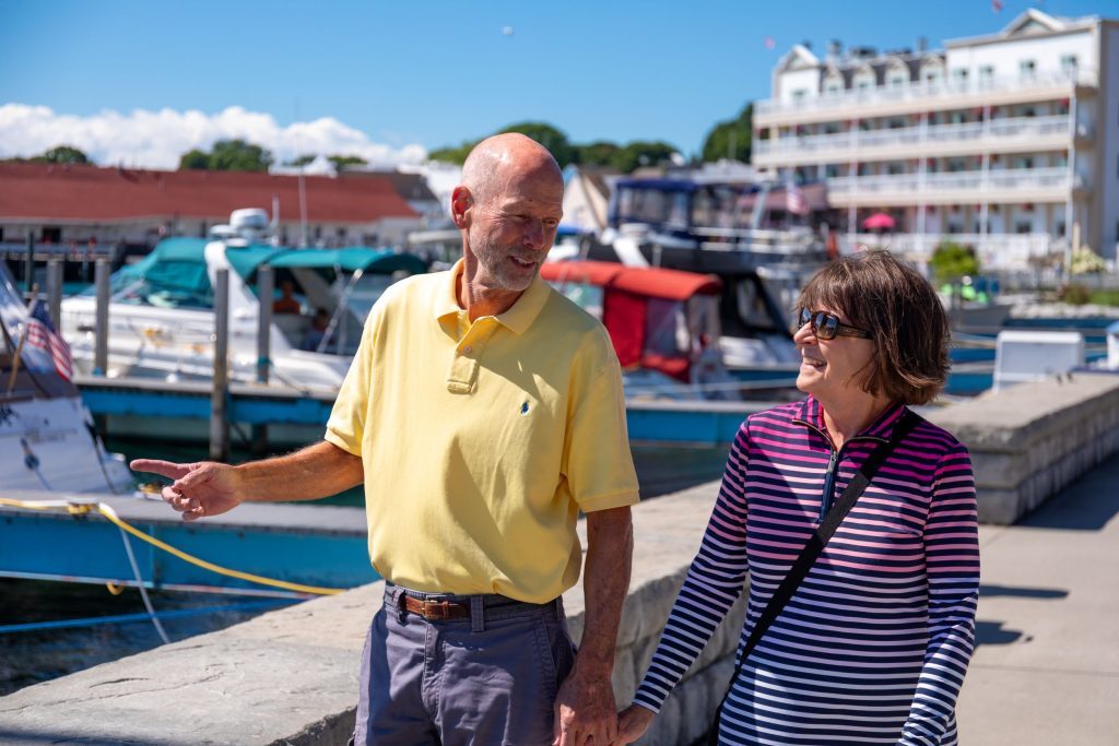 A couple walks along the waterfront past boats in Mackinac Island’s harbor.