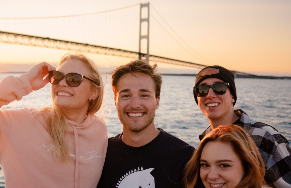 A group of four young people wearing Mackinac Island clothing smile for the camera on a boat near the Mackinac Bridge