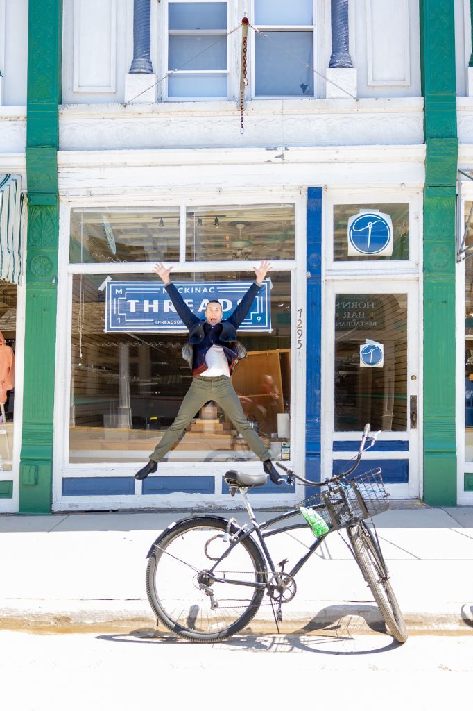 A man jumps in front of Mackinac Island’s Threads of Mackinac storefront window with a bicycle in the foreground 