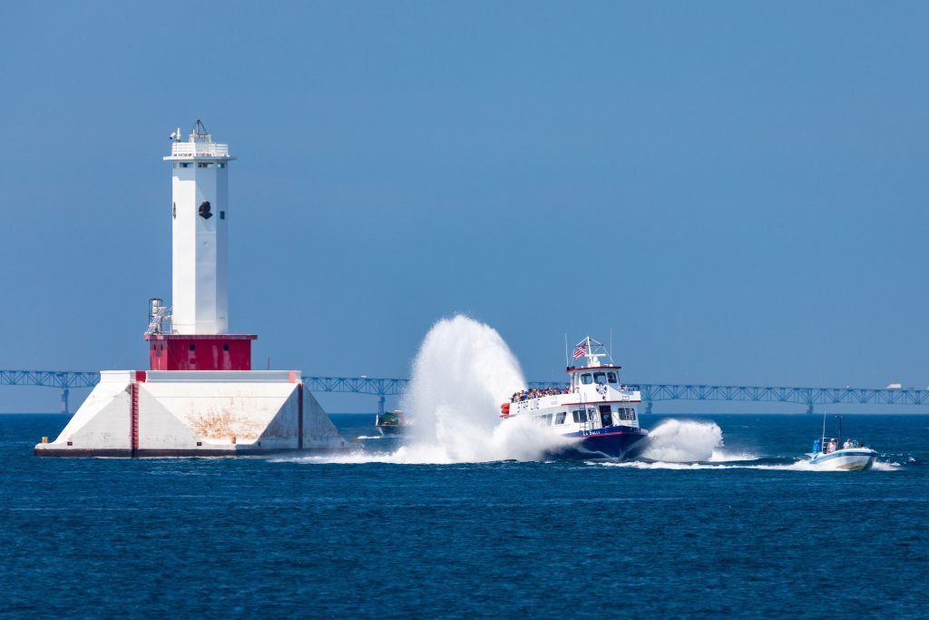 A Mackinac Island ferry boat kicks up a tail of water as it passes a light station with the Mackinac Bridge in the background