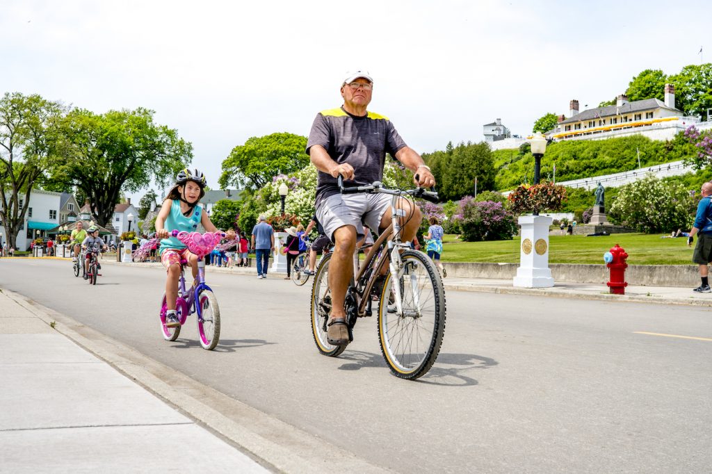 A man and a young girl ride bicycles down the street on Mackinac Island in front of historic Fort Mackinac