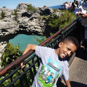 A young boy smiles in front of Mackinac Island's Arch Rock as he holds the railing of the viewing platform