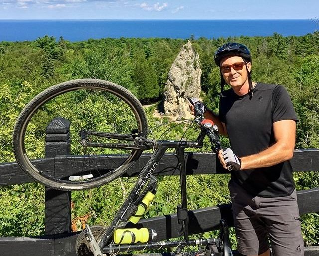 A bicyclist wearing a helmet poses with his bike with Mackinac Island's Sugar Loaf in the background