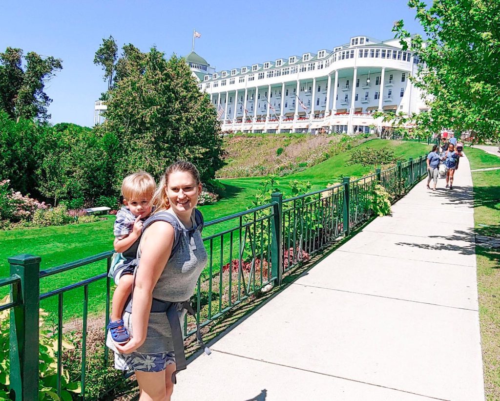 A young woman with a child on her back smiles on a sidewalk in front of Mackinac Island's Grand Hotel