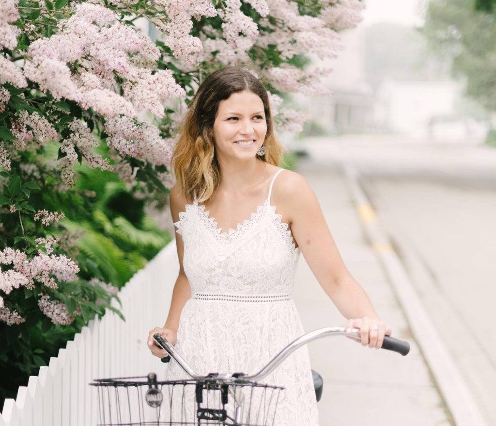 A young woman stands holding the handlebars of a bicycle on a Mackinac Island sidewalk with lilacs overhead