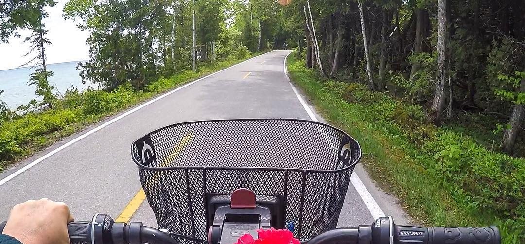 Bicycle handlebars and a basket on a Mackinac Island road lined with trees and water