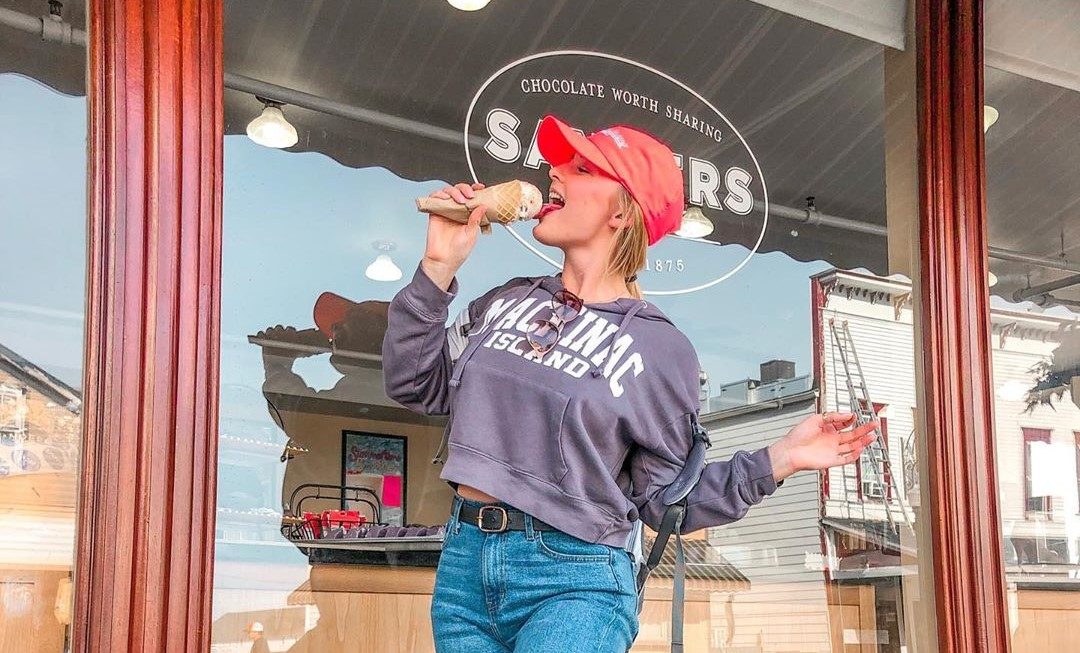 A young woman eats ice cream while wearing a Mackinac Island sweatshirt in front of Sander's Fudge shop