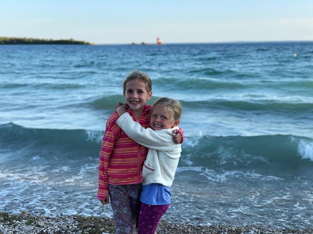 Two young girls hug each other by the water on the beach at Mackinac Island’s Windermere Point with Round Island Lighthouse in the background