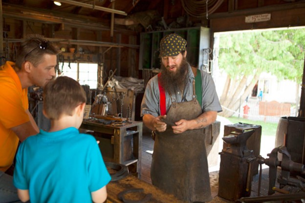 A blacksmith explains his metal forging process to a father and son on Mackinac Island