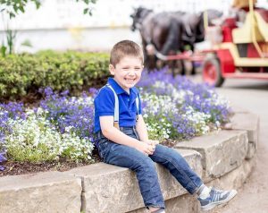 A smiling young boy sits on a wall with flowers and a horse-drawn carriage behind him on Mackinac Island