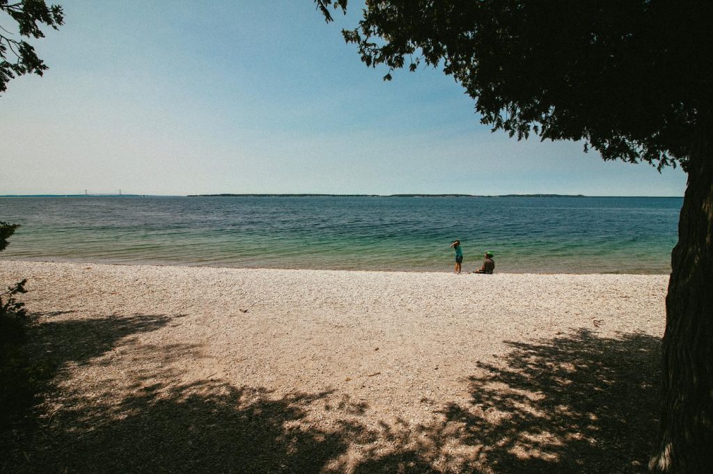 Two human figures are silhouetted on a rocky Mackinac Island beach with beautiful blue water