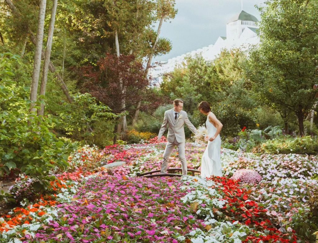 A bride and groom walk over a bridge in a gorgeous flower garden near Mackinac Island's Grand Hotel