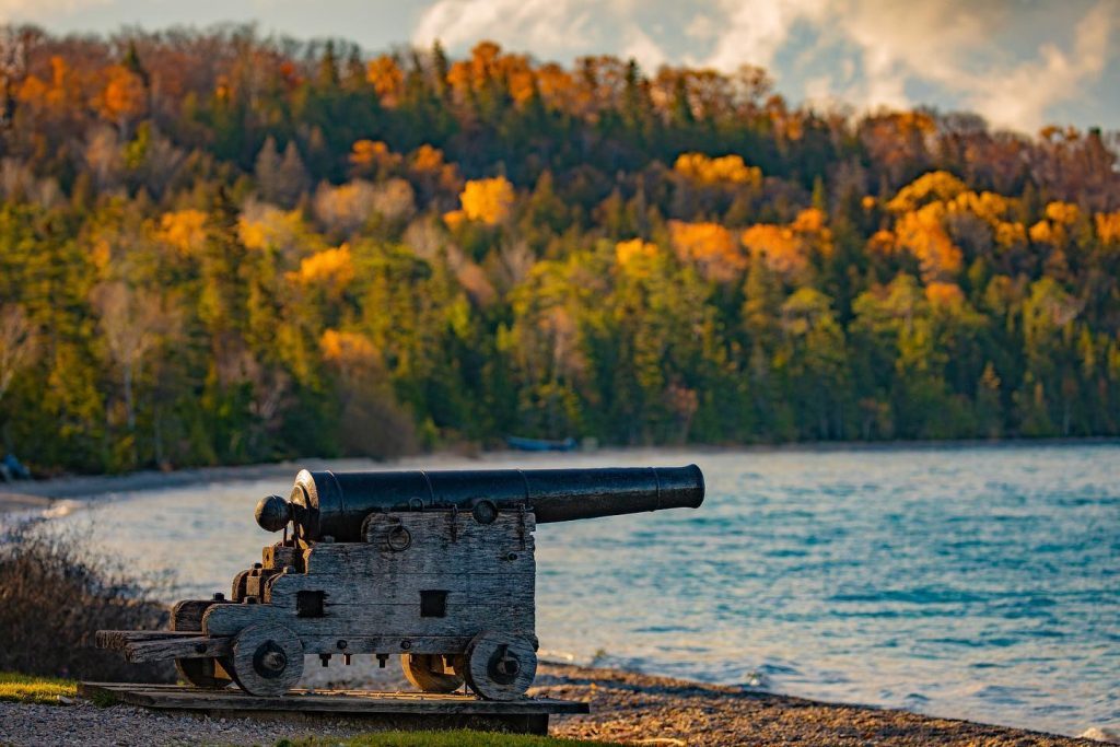 A cannon points over the water from the beach at Mackinac Island’s British Landing with fall color in the background