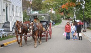 Women with shopping bags stand on the side of a Mackinac Island road as a horse-drawn carriage passes by in fall