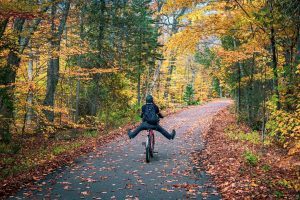 A man takes his feet off the pedals of a bicycle while riding down a paved path in the woods of Mackinac Island with leaves on the ground in fall
