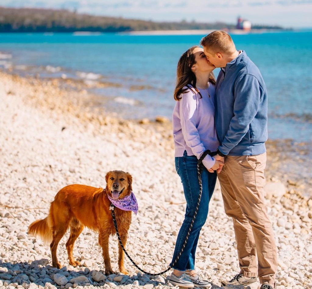 A couple kisses while holding a dog on a leash on the rocky beach of Mackinac Island’s Windermere Point with Round Island Lighthouse in the background