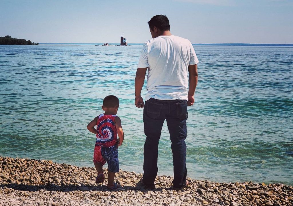 A father and young son stand by the water on the rocky beach at Mackinac Island’s Windermere Point with Round Island Lighthouse in the background 