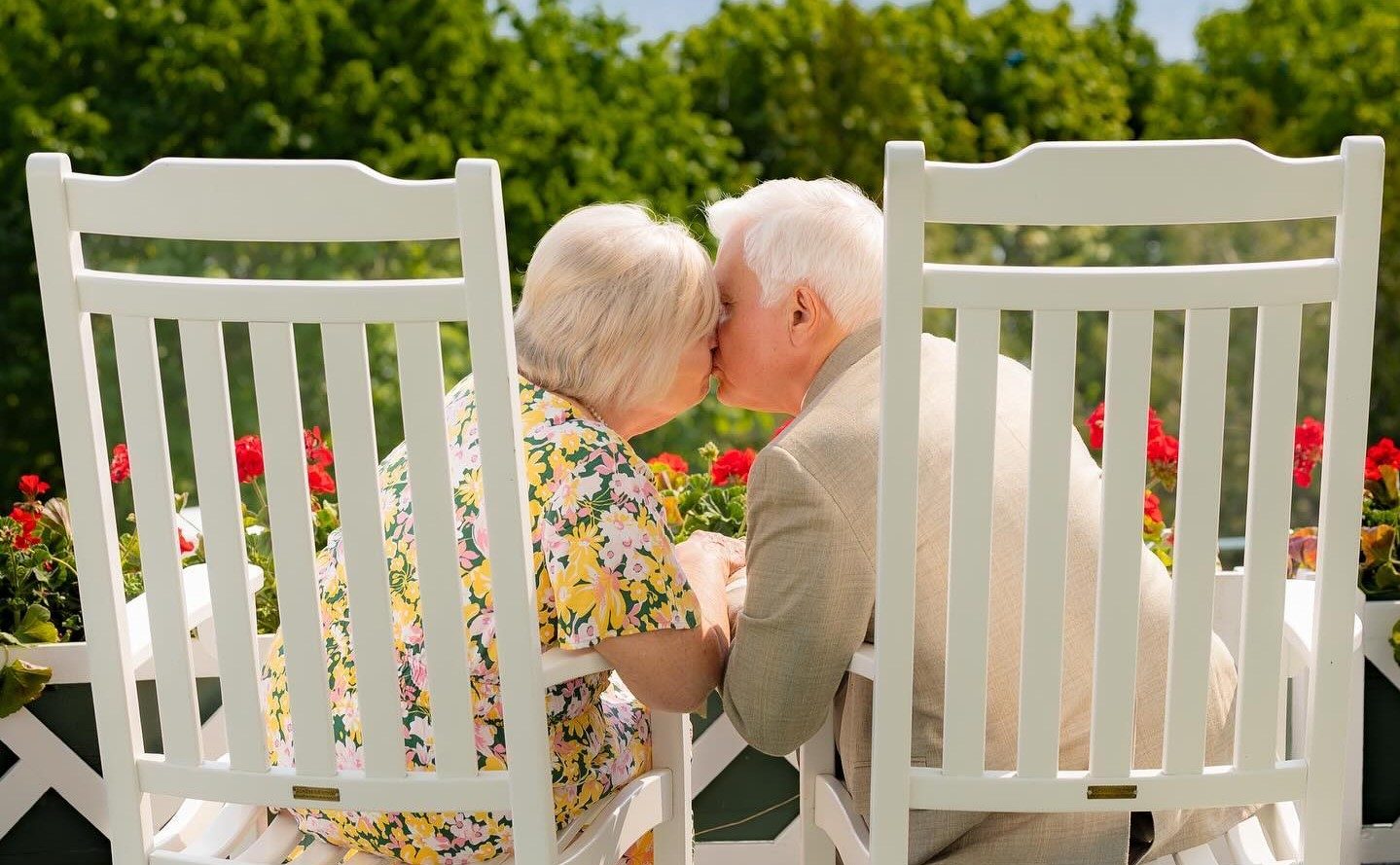 An older couple leans in for a kiss while sitting in white Adirondack chairs on the porch of Mackinac Island’s Grand Hotel