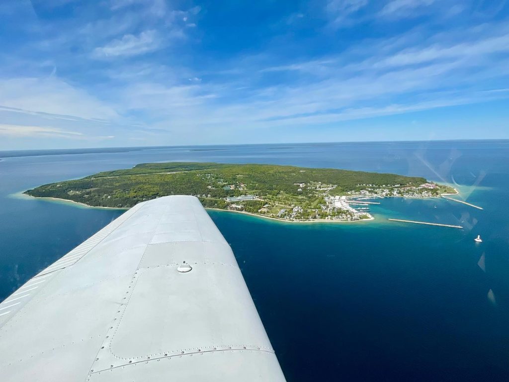 The wing of an airplane points to Mackinac Island down below surrounded by water in the middle of the Straits of Mackinac