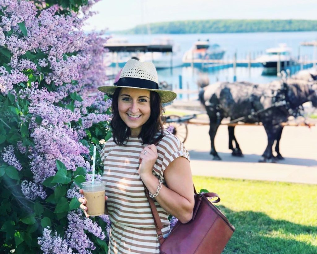 A young woman wearing a hat poses by a lilac bush in Marquette Park in front of horses and the Mackinac Island marina
