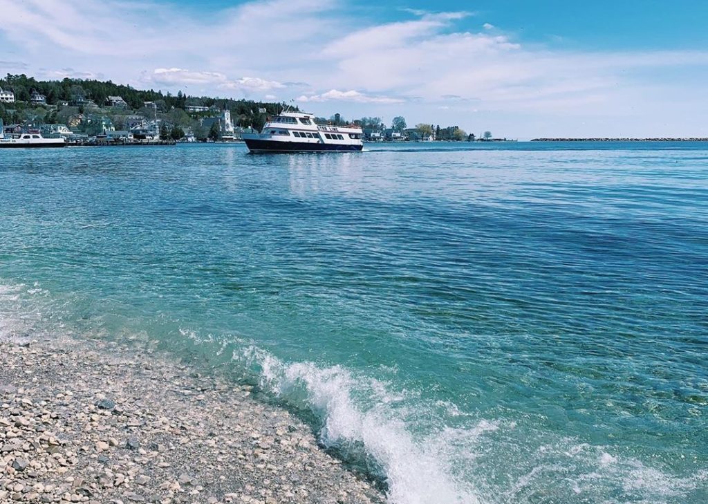 A Mackinac Island ferry boat approaches the dock on a sunny day as seen from the rocky beach at Windermere Point