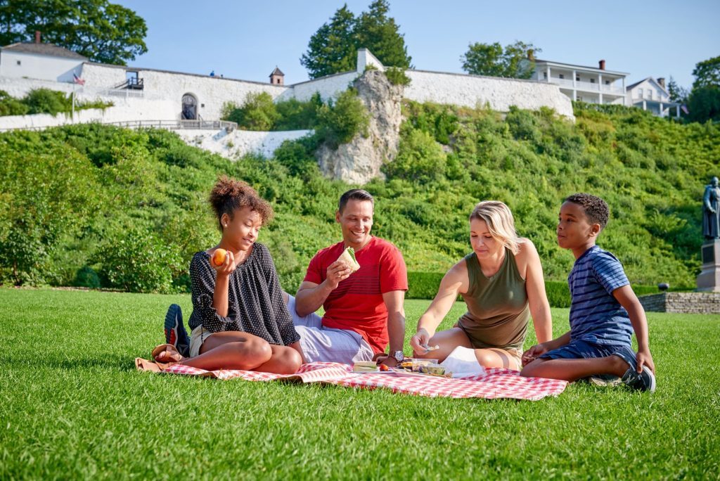 A family of four has a picnic on a blanket on the grass in Marquette Park below Fort Mackinac