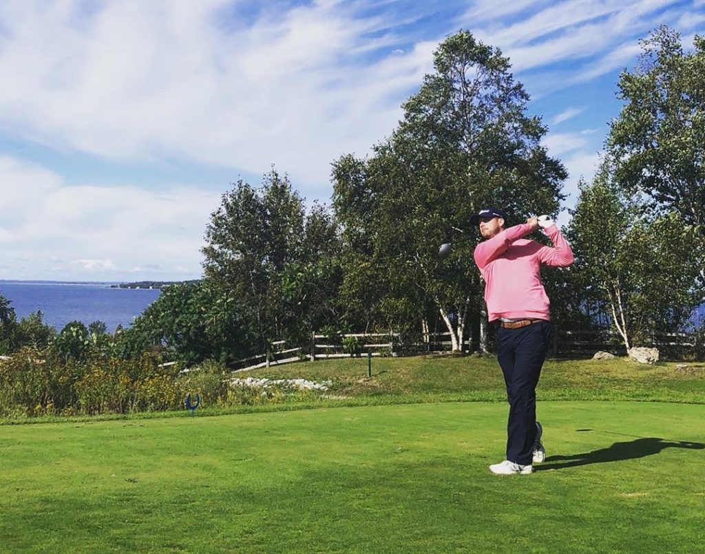 A man in a pink sweater watches the flight of his golf ball after swinging on a tee at Mackinac Island's Jewel Golf Club at Grand Hotel