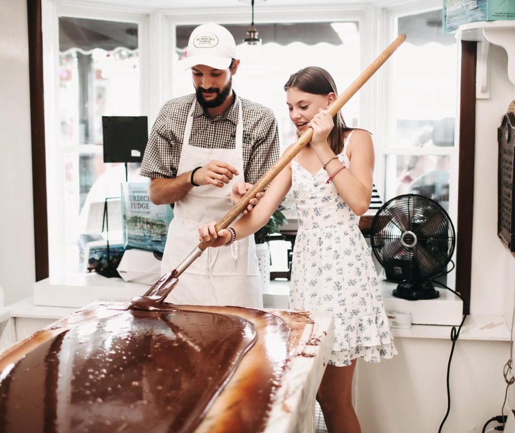 A young woman tries her hand at making fudge as a fudge maker watches in a Mackinac Island fudge shop