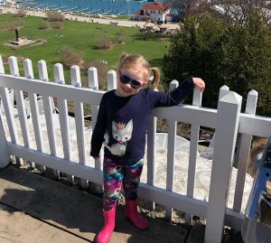 A young girl stands by a white picket fence at Fort Mackinac overlooking Marquette Park and the Mackinac Island marina