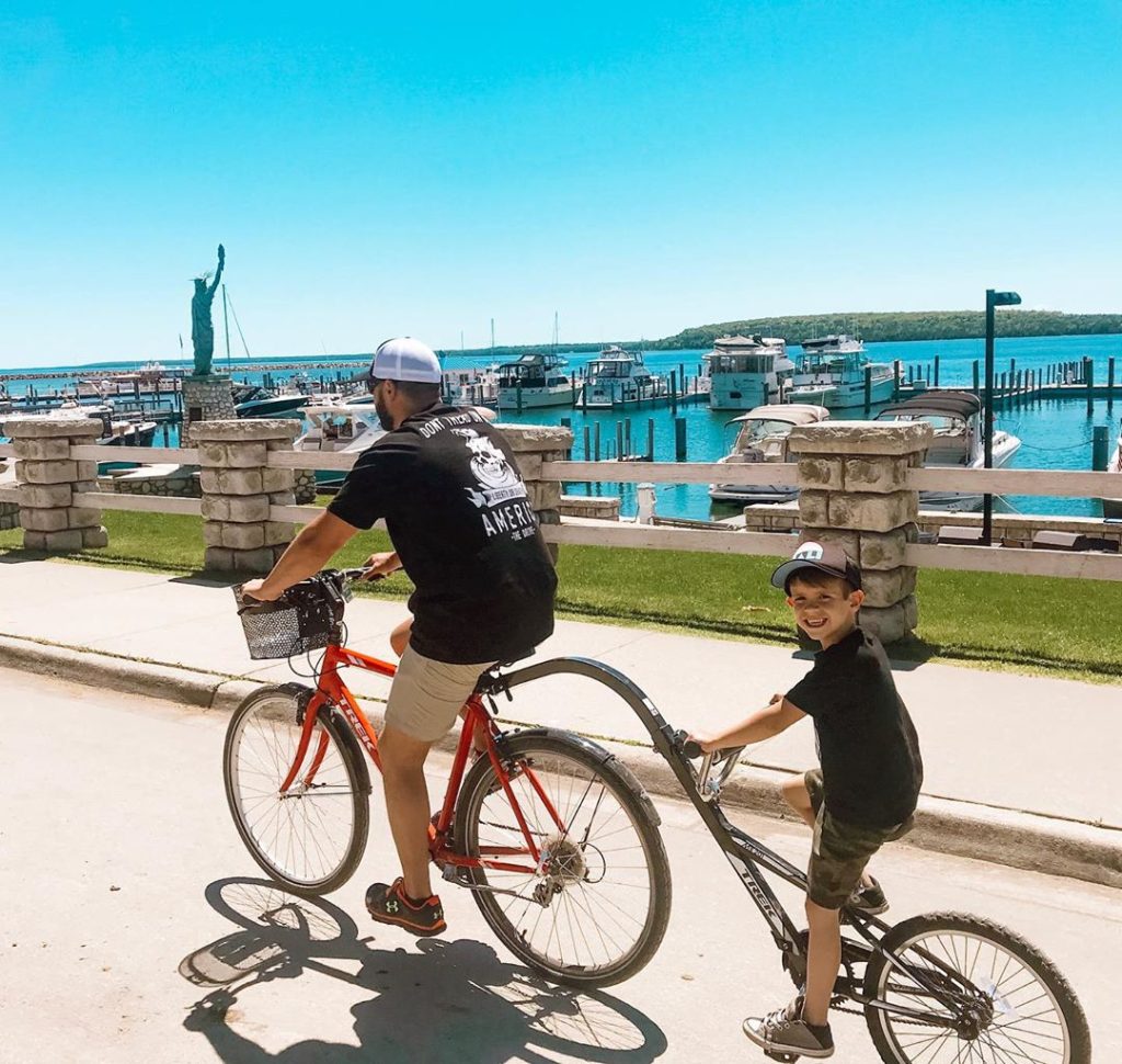 A young boy pedals a tagalong behind has dad's bicycle on a road along the Mackinac Island marina