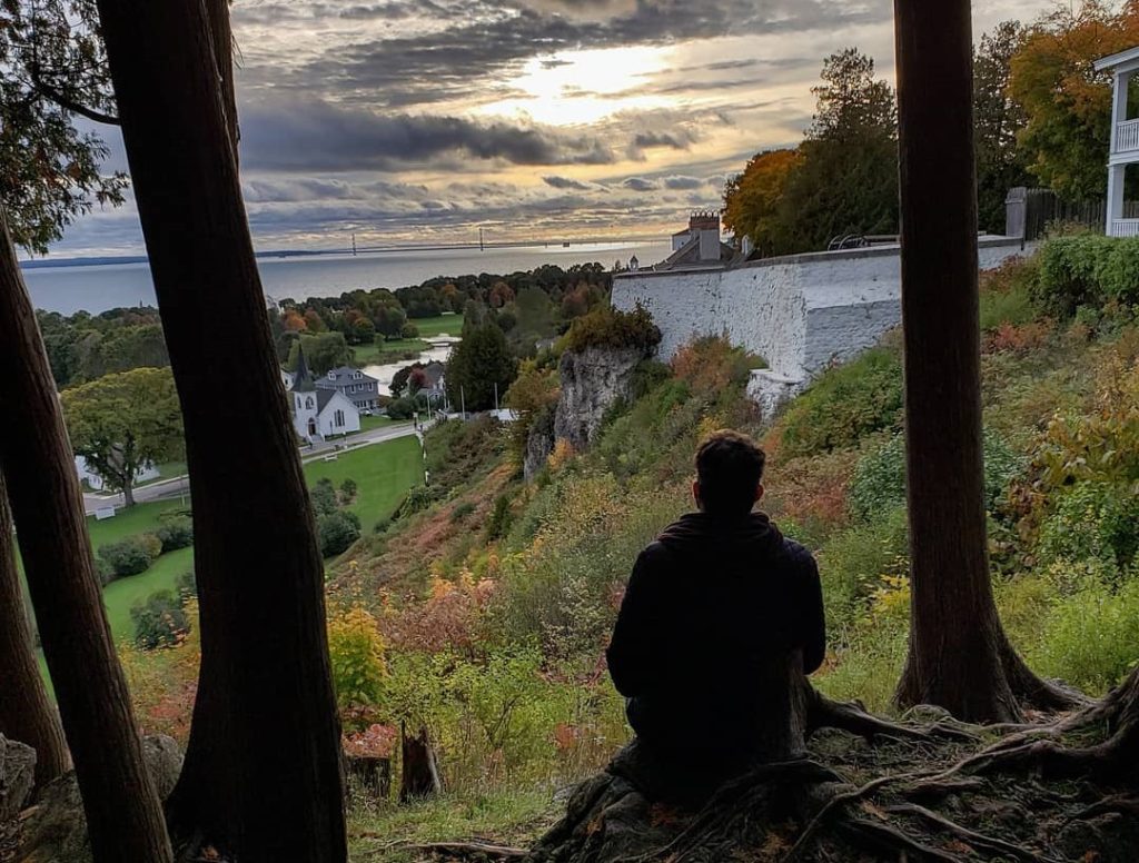 A hiker silhouetted by the setting sun sits on a bluff overlooking downtown Mackinac Island in fall